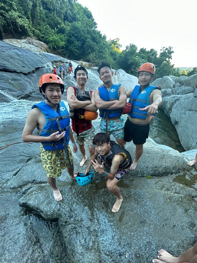 Five friends in helmets and life jackets posing on rocks beside a river