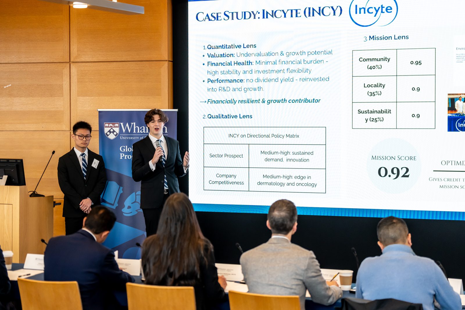 Two students presenting beside a projected slide titled Case Study: Incyte, with judges seated in the foreground